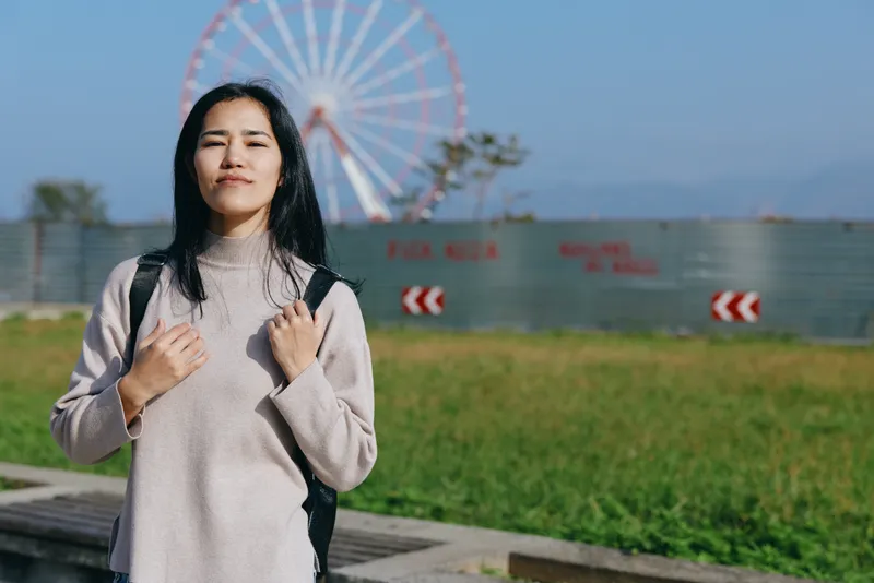 Woman with backpack in outdoor park beside a ferris wheel, casual attire enjoying sunny day and urban scenery