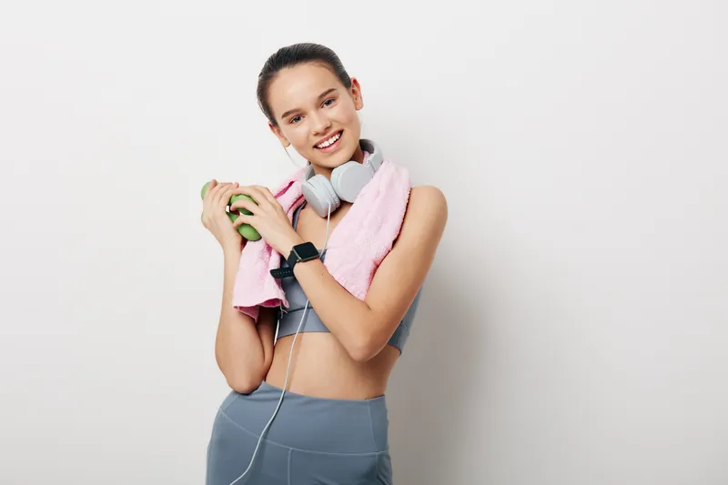 Fitness woman in athletic wear with a towel and headphones after a workout, smiling and holding a water bottle against a plain wall, showcasing energy, health and sport practice.