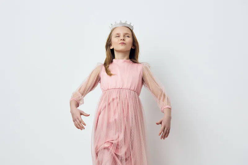 Young girl in a pink dress with a crown, exuding confidence and joy against a minimalist white background, perfect for concepts of childhood dreams and royal themes
