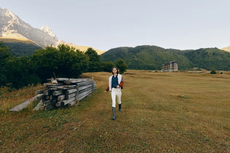 Man walking in a meadow field with distant mountains and countryside building, wearing boots and casual jacket, outdoor hiking scene, nature landscape at warm sunlight and peaceful solitude.