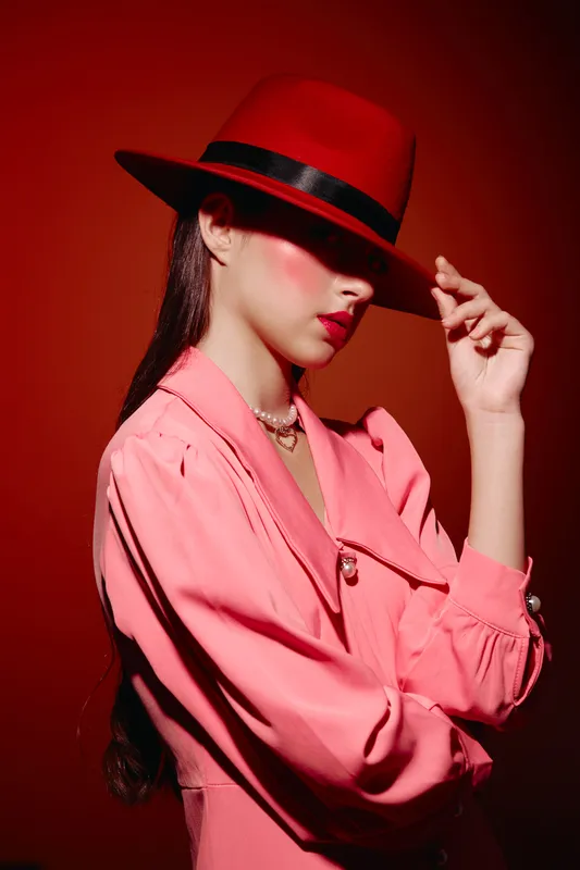 Fashionable woman in a red hat hides her eyes with the brim. She wears a pink jacket with pearl necklace, styled in creative beauty studio lighting on red background.