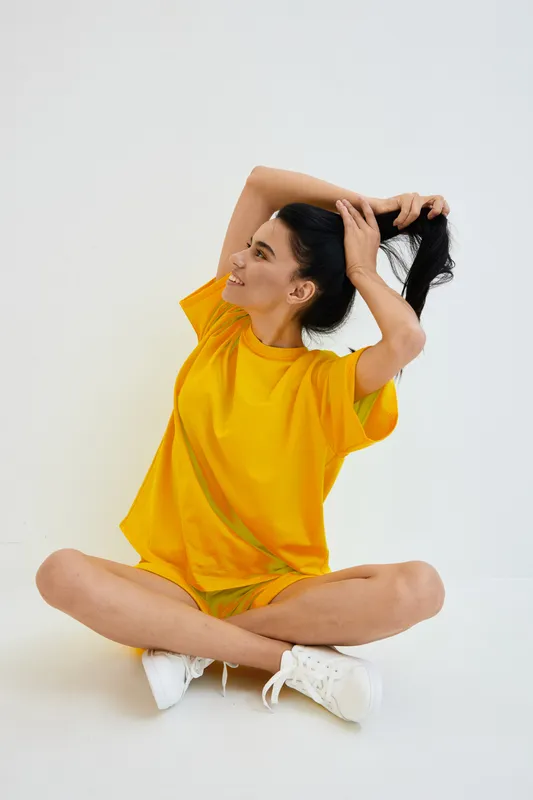 Young woman sitting cross-legged in yellow casual dress, smiling joyfully while tying her hair, isolated on white background with bright studio lighting.
