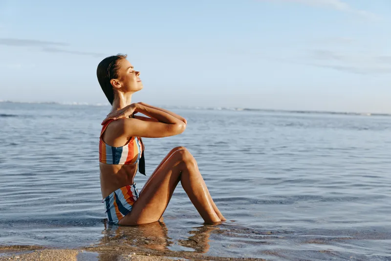Woman girl on beach by ocean wearing swimsuit, sitting in shallow water, relaxation pose, calm gaze upward, gentle waves behind, summer outdoors lifestyle