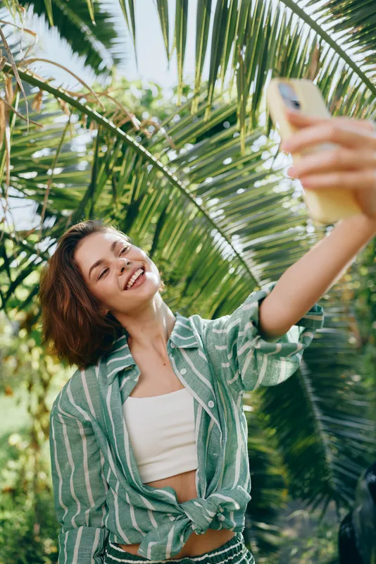 Young woman taking a selfie outdoors amid lush palm leaves, smiling warmly as she captures a bright, sunlit moment.