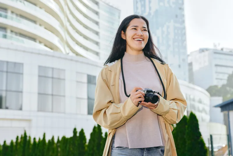 Woman with camera in hand, wearing a beige jacket and sweater, smiling outdoors in a modern city setting, urban photography and lifestyle shoot during sunny afternoon, travel vibe