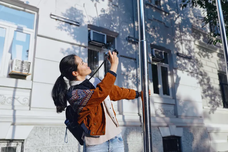 Woman photographer captures street scene with camera and lens beside a classic building, outdoor urban setting, candid action, precise focus on photography technique, composition, light, and street