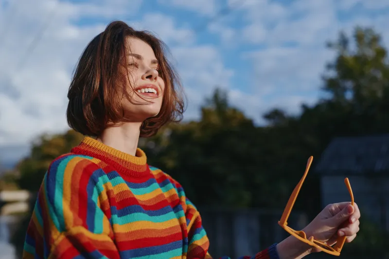 Woman smiling in a colorful striped sweater outdoors, young adult with short hair holding sunglasses, casual outfit, joyful expression and sunlit lifestyle portrait in a backyard.