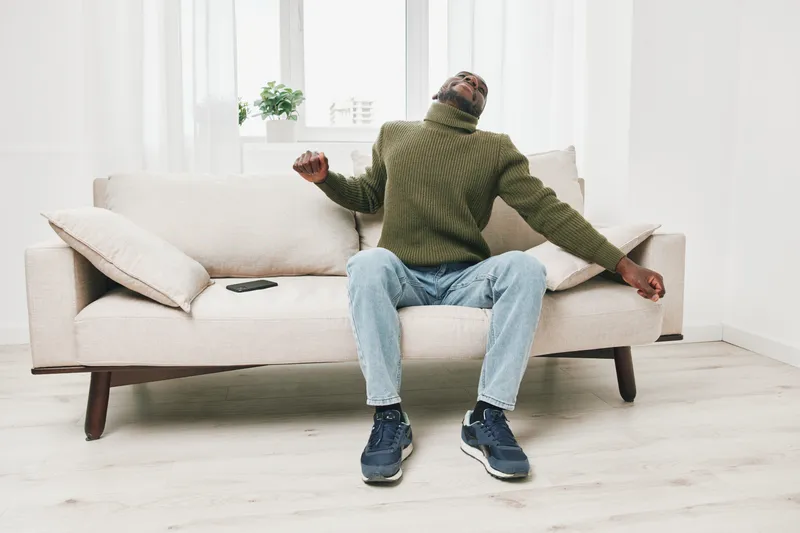 Young Black man relaxing on a beige couch in a light filled room, wearing a green turtleneck sweater and blue jeans, expressing calmness and comfort