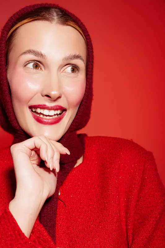 Smiling woman in red sweater and hood looks to the side with bright lipstick and glowing skin. Close-up portrait on red background. Concept of happiness, fashion, and beauty.