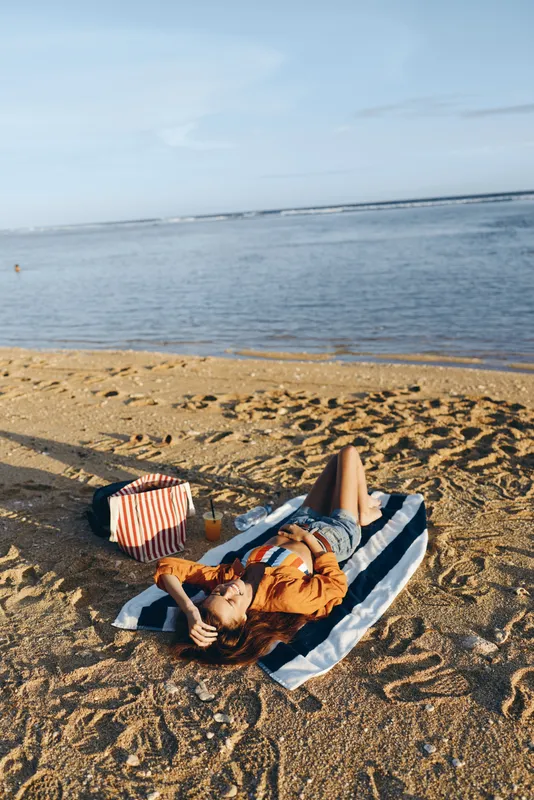 Young woman relaxing on striped towel on sandy beach near calm sea under clear sky enjoying peaceful sunny day outdoors with bag and drink nearby