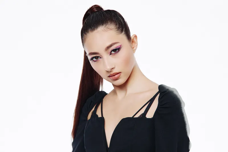 Young woman with creative cat-eye eyeliner looks confidently at camera against a white background. The photo emphasizes fashion, beauty, and modern makeup style.