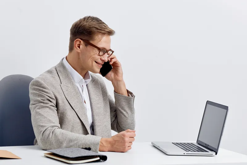 Professional man in glasses talking on mobile phone at office desk with laptop and notepad on white background, business communication, workplace, corporate, modern office environment.