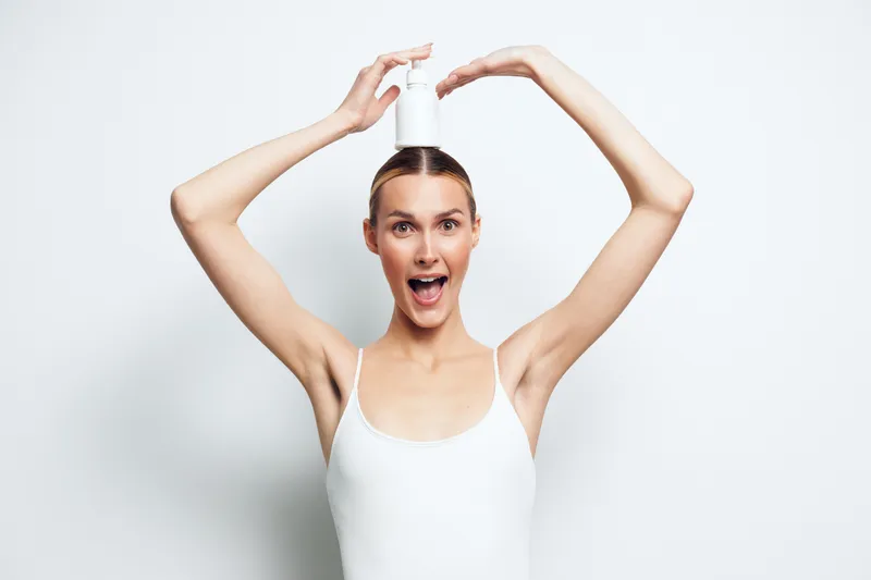Young woman with clear skin holding a white lotion bottle above her head, wearing a white tank top, smiling with open mouth, bright studio background, skincare concept