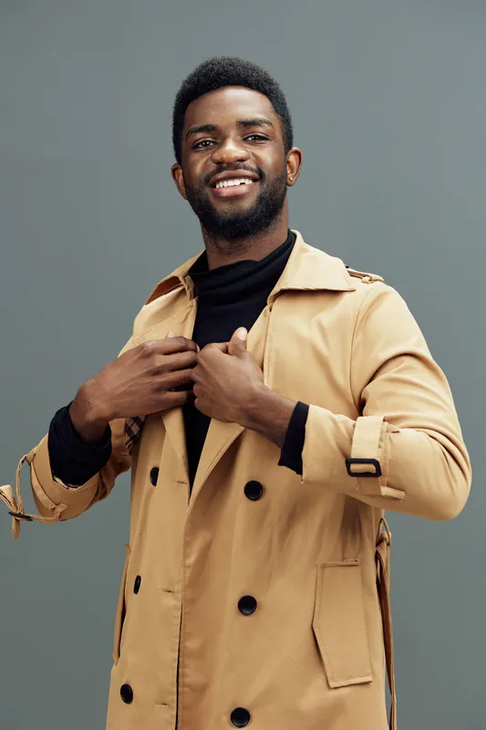 Stylish young African man in a beige trench coat smiling confidently against a minimalist gray background, showcasing joy and fashion forward attire