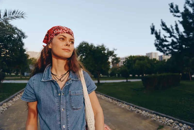 lifestyle woman in modern boho-western denim street style stands confidently outdoors with film color tones, wearing a red bandana and layered necklaces under natural sunlight