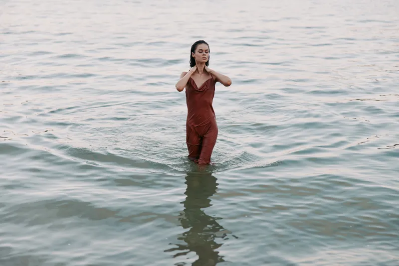 A woman stands waist-deep in calm sea water, wearing a red dress. The soft light and gentle waves create a serene, intimate moment of quiet solitude.