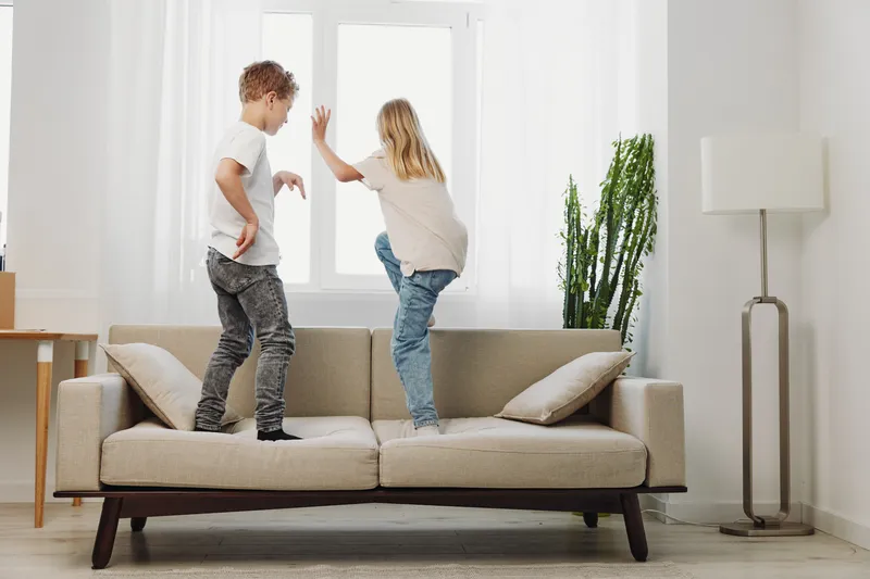 Two playful children jumping on a beige sofa in a bright living room with modern decor and natural light. Concept of childhood fun, energy, and family lifestyle at home.