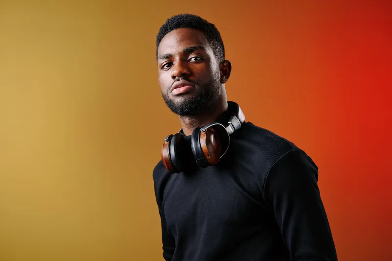 Portrait of a confident young African man with headphones, wearing a stylish black sweater against a vibrant orange yellow gradient background