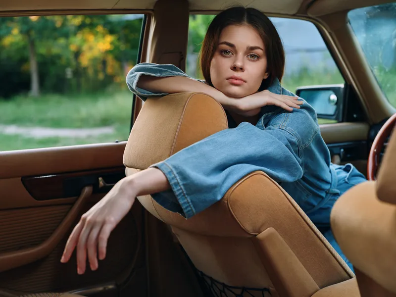 Young woman in denim jacket leaning casually in a vintage car, evoking a serene summer vibe with natural light and green background.