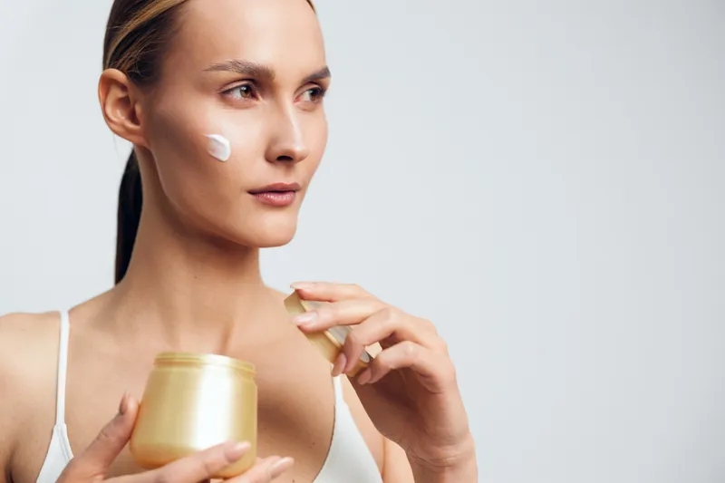 A young woman applies cream to her cheek while holding a jar of skincare product. The bright neutral background highlights her natural complexion and beauty routine concept.
