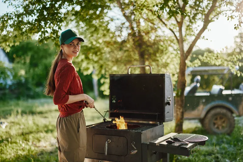 Smiling woman grilling outdoors in summer, wearing casual attire and a cap, with a vibrant green background and warm sunlight illuminating the scene