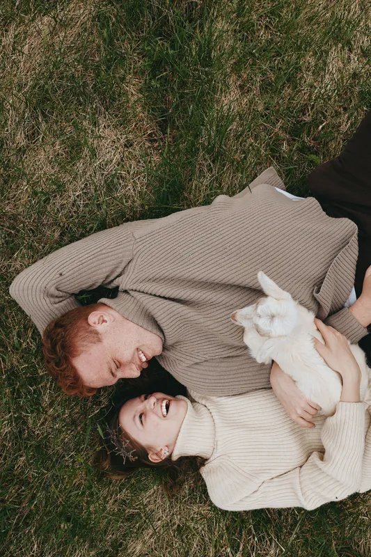 A couple lies on soft grass sharing a warm moment, wearing knitted sweaters, while a playful goat kid rests beside them in a cozy countryside scene.