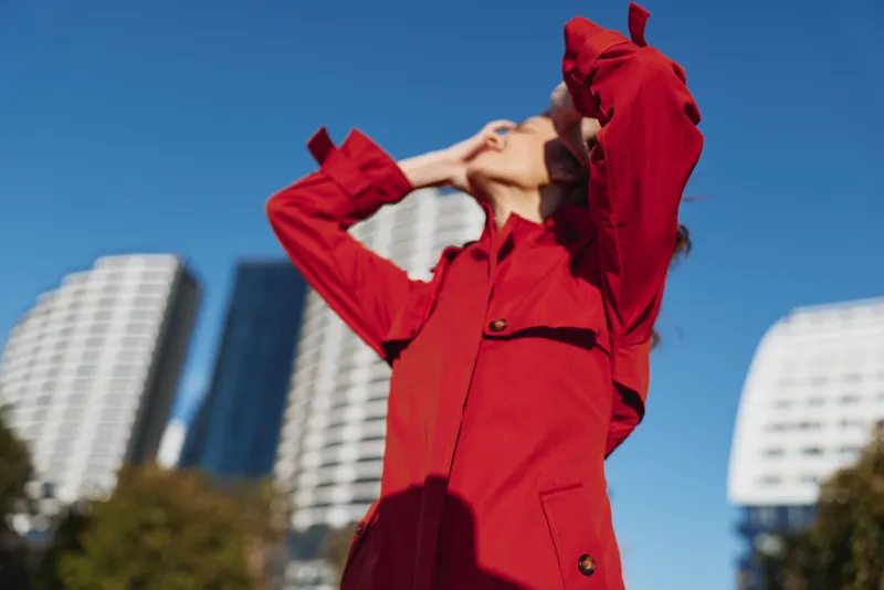 Young woman in a red coat enjoys a sunny day outdoors with her hands raised to her face against a clear blue sky and modern buildings background.