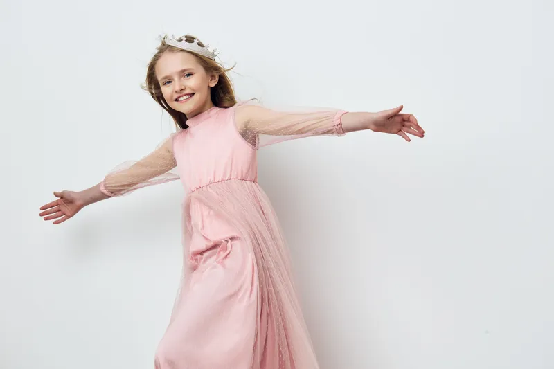 A joyful young girl in a pink dress with flowing sleeves, posing playfully against a minimalist white background, capturing innocence and happiness