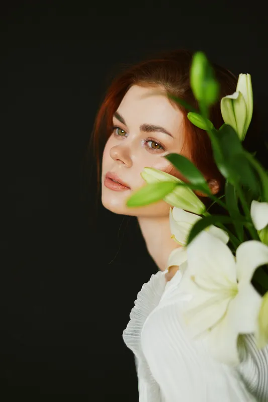Young woman with red hair holding white lilies, wearing a light sweater, showing a calm expression against a dark background Fresh floral concept