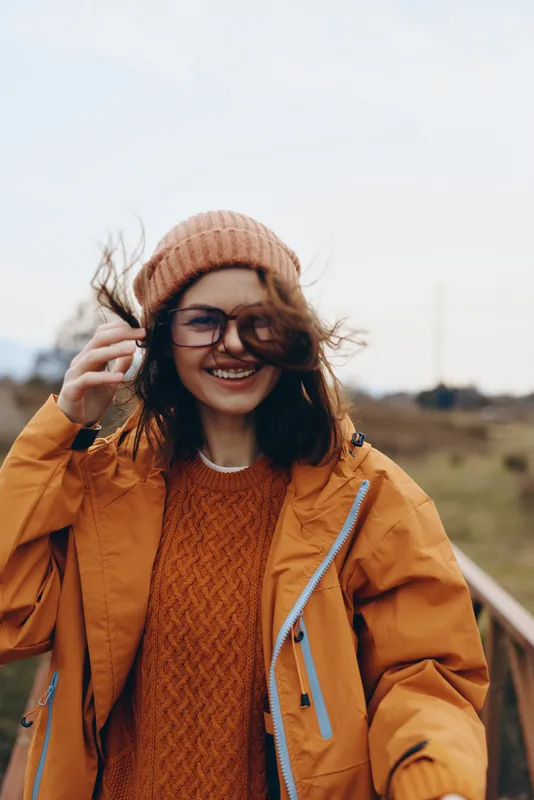 Woman lifestyle outdoors smiling while talking on phone, wind lifts hair, she wears beanie, glasses, orange jacket and knit sweater, casual autumn street style on a rural path in daylight