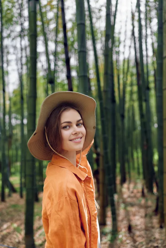 Woman smiling in a straw hat and orange jacket, portrait in a bamboo forest. Outdoor nature scene with soft light, relaxed travel vibe and peaceful candid expression on a young model.