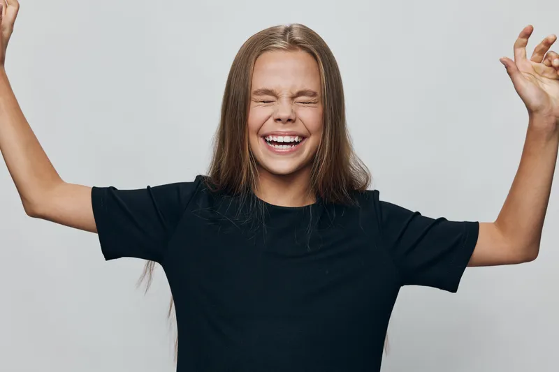 Girl with long hair wearing black shirt smiling with eyes closed and raised arms against gray background for happiness joy and excitement concept.