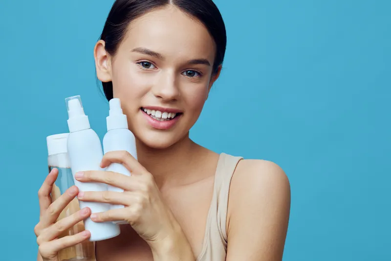 Young woman holding skincare products, showcasing healthy skin and a natural beauty routine against a vibrant blue background, ideal for wellness and skincare concepts