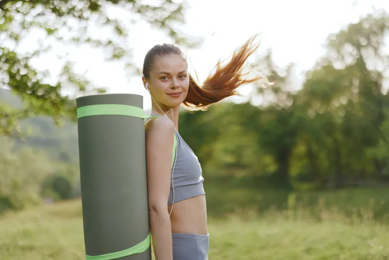 Young woman carrying a yoga mat outdoors, showcasing a healthy lifestyle in a serene natural environment filled with greenery