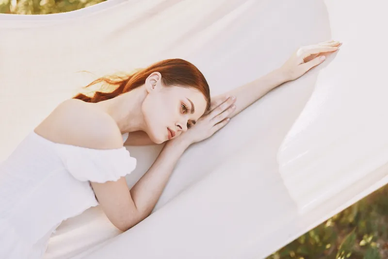 Young woman in a white dress resting on soft fabric, expressing calm emotions in a serene outdoor setting, ideal for wellness and relaxation themes