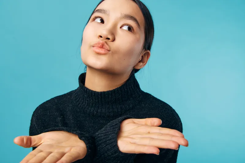 Young woman with dark hair wearing black sweater posing with hands open and looking up against blue background. Female model expressing doubt or curiosity in studio portrait.