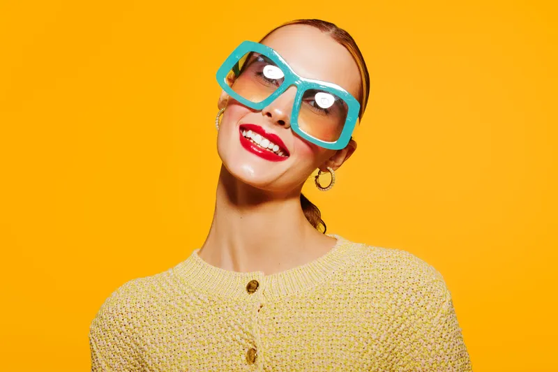 Smiling woman with large blue glasses and red lipstick poses against bright yellow background. She wears a textured yellow sweater and gold hoop earrings. Concept of fashion and joy.