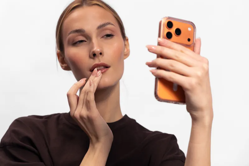 Young woman with clear skin holds orange smartphone looking at screen, applying lip balm. Isolated on white background. Concept for skincare, beauty, and digital lifestyle.