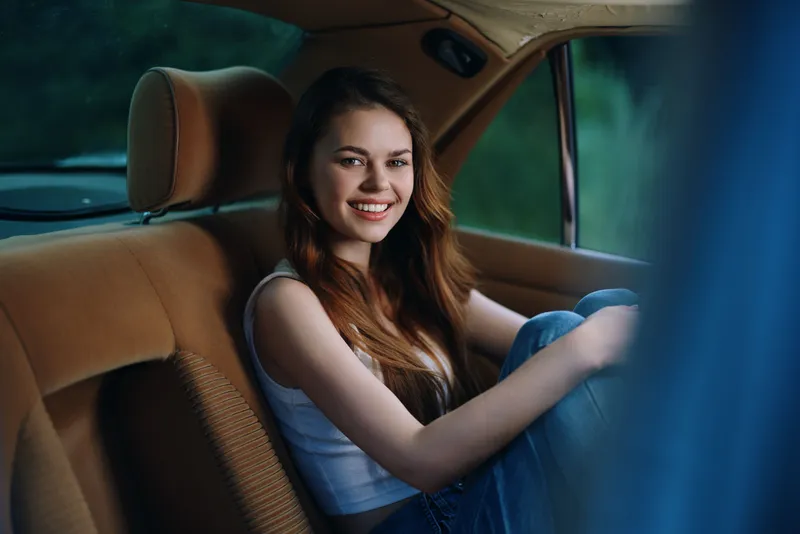 Young woman smiling in a vintage car interior, showcasing casual fashion and joyful emotions in a serene outdoor setting.