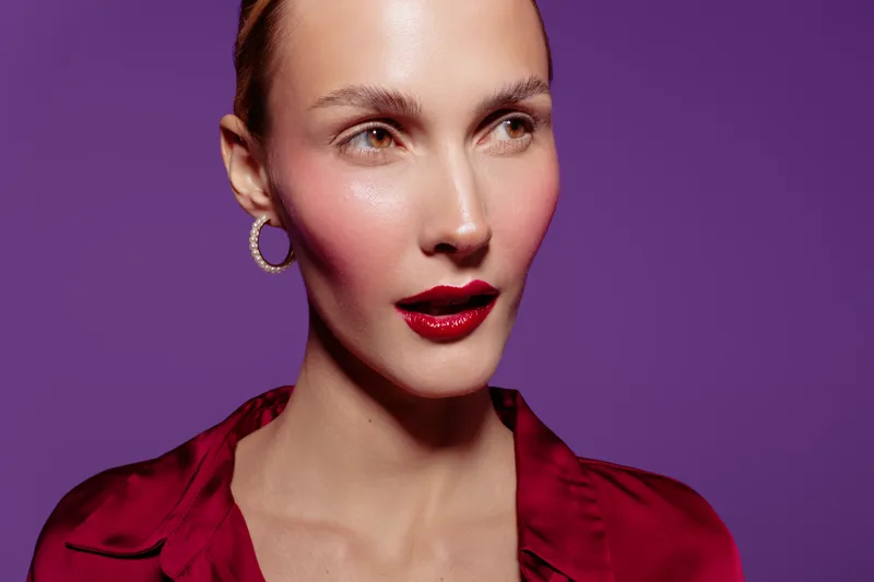 Close-up portrait of a woman with red lipstick and blush wearing a red blouse and hoop earrings on purple background. Concept of beauty, fashion, and makeup.