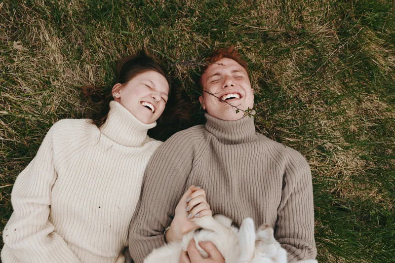 A joyful couple lies on the grass in cozy sweaters, sharing a laugh while cuddling a fluffy animal, capturing a candid lifestyle moment of affection, comfort, and carefree companionship