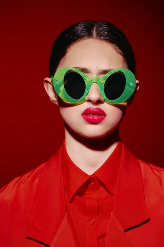 Portrait of a young woman wearing bright green sunglasses and red lipstick. She wears a vibrant red blazer and shirt. High fashion concept with strong color contrast and beauty creativity.