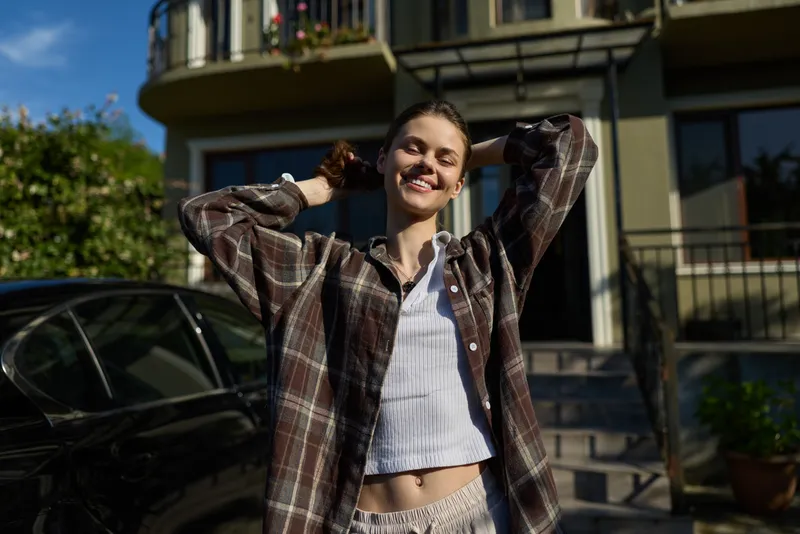A cheerful woman in a plaid overshirt stands outdoors in daytime, smiling confidently, casual layered outfit, city street backdrop, bright natural light, lifestyle portrait