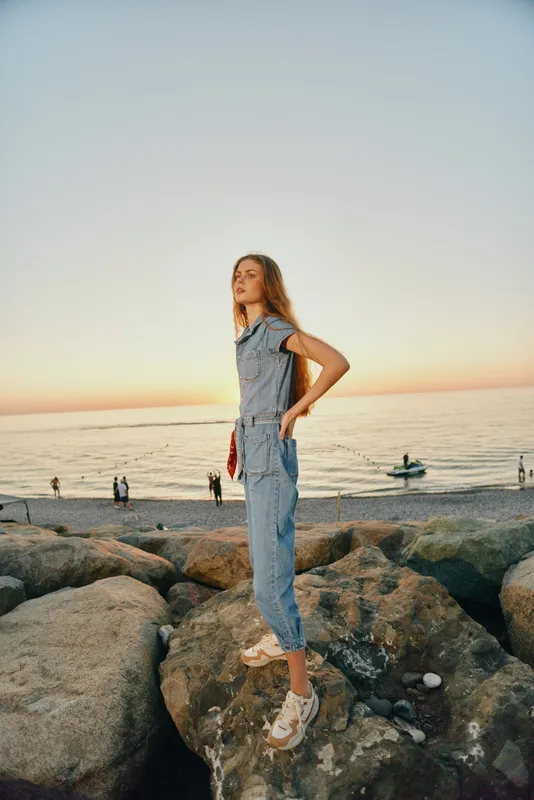 Lifestyle woman in modern boho-western denim street style stands confidently on rocky shore during golden hour with warm film color tones.