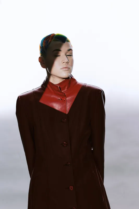 woman with colorful braided hair wearing a dark red leather jacket standing with eyes closed against bright white background in dramatic lighting