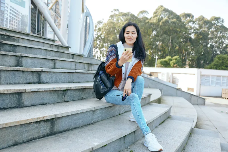 Casual outdoor scene with a woman sitting on urban stairs, smiling at a smartphone while resting a backpack beside her, showcasing fashion and relaxed lifestyle on a sunny day.