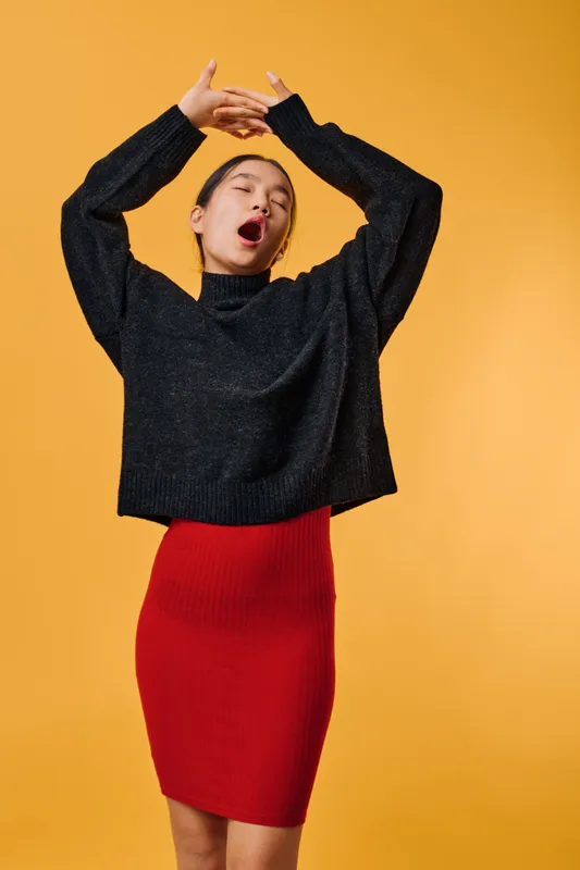 Woman wearing black sweater and red skirt yawning and stretching arms up on bright yellow background, sleepy expression and casual outfit for indoor lifestyle and relaxation.