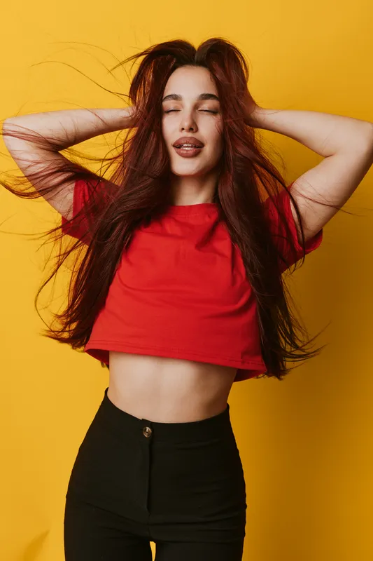 Young woman with long hair and closed eyes posing with hands in hair wearing red crop top and black pants on yellow background in studio. Fashion, beauty and casual style portrait.