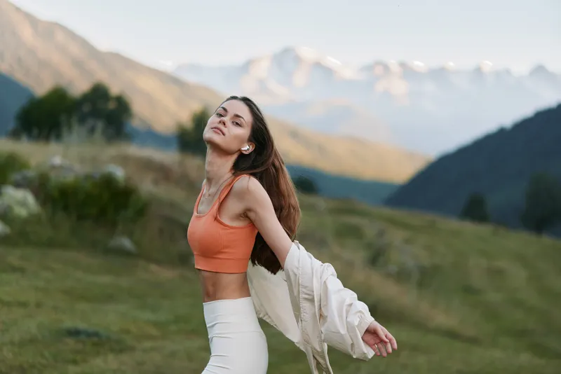 A cheerful woman poses outdoors in a mountainous landscape. She wears an orange crop top and white pants, with a light jacket draped over her shoulder, enjoying a sunny, fresh air moment.