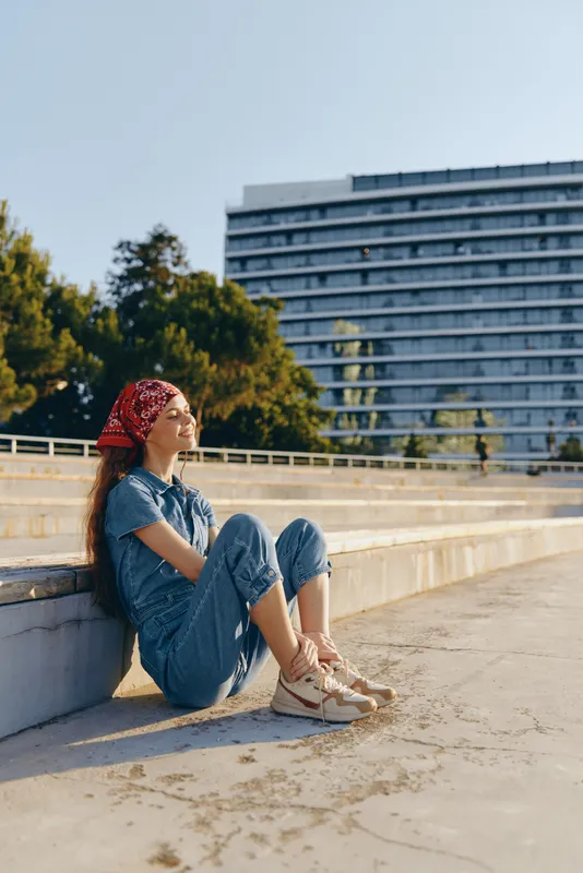 relaxed woman sitting outdoors in casual clothes enjoying sunlight near urban building on clear day with green trees and blue sky background